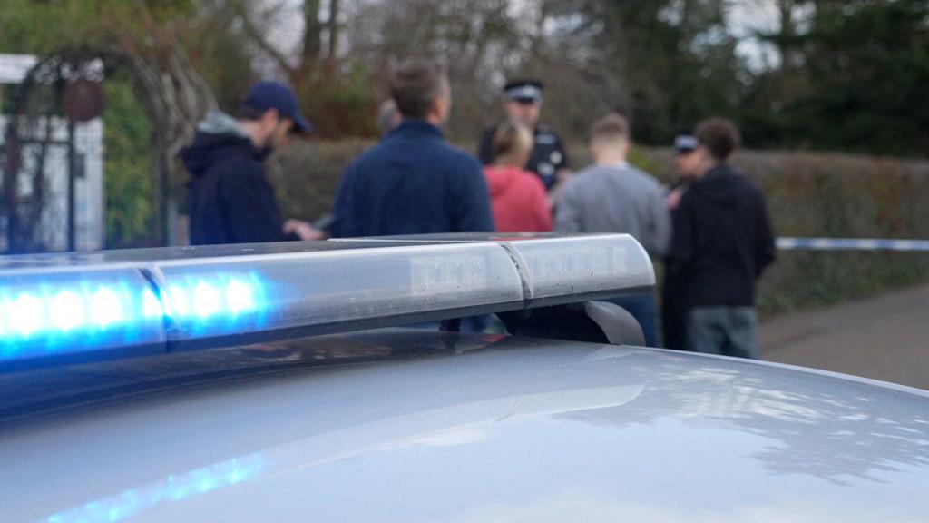 In the foreground in focus is the top of a police car with a blue light on. Behind out of focus are a crowd of people and a uniformed police officer in black.