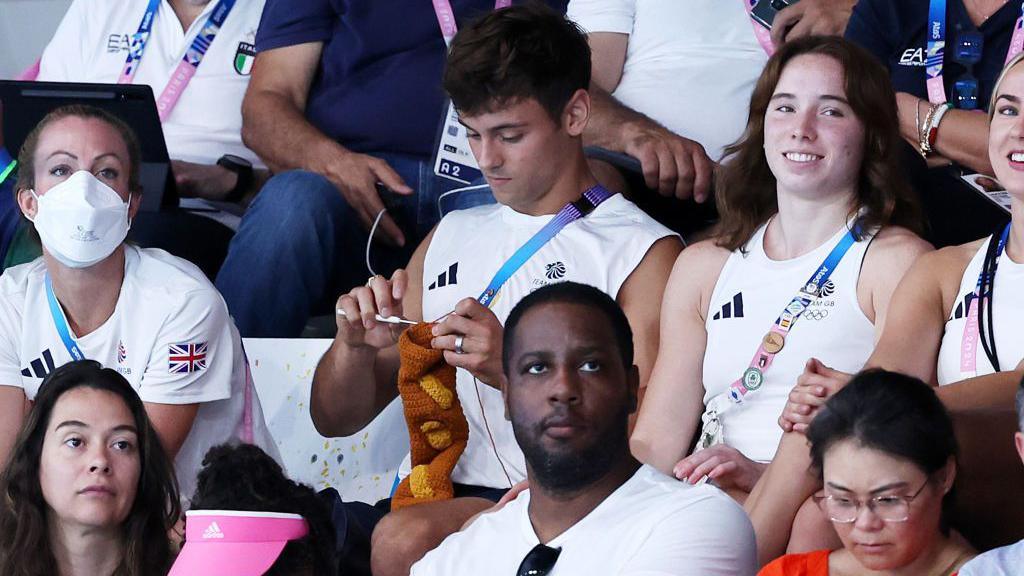 Diver Tom Daley sitting in a crowd at the 2024 Olympic Games knitting a brown sock or sleeve. 