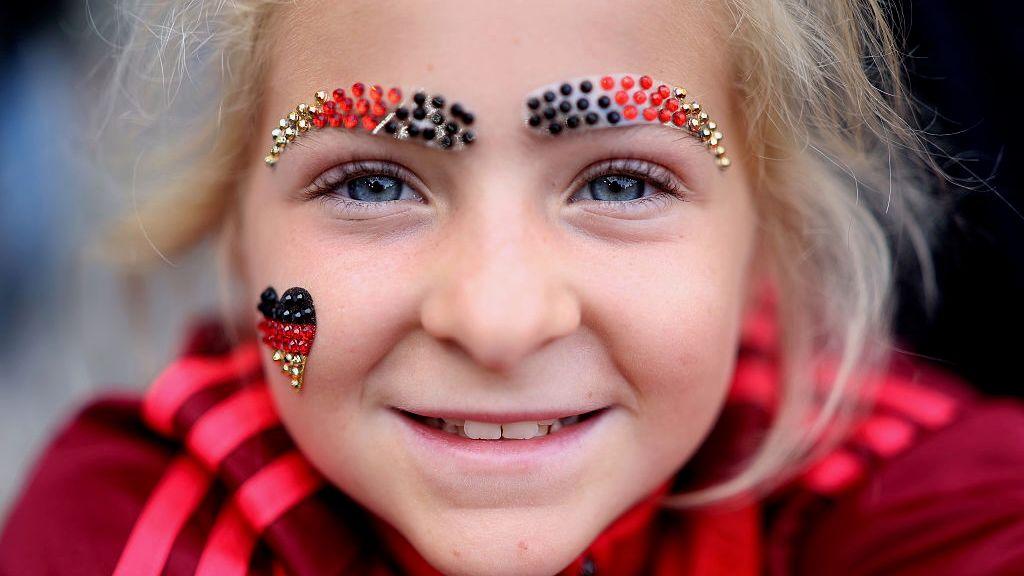 A young girl with blonde hair and blue eyes smiles to the camera in a close up shot. She is wearing sparkly eyebrows made out of beads in the colours of the German flag and a little heart on her right cheek.