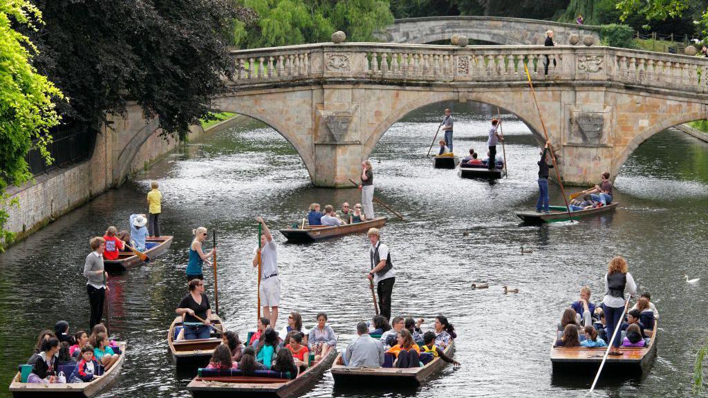 There are several punts on the River Cam with people sitting in them. There are two bridges over the river and trees on either side.
