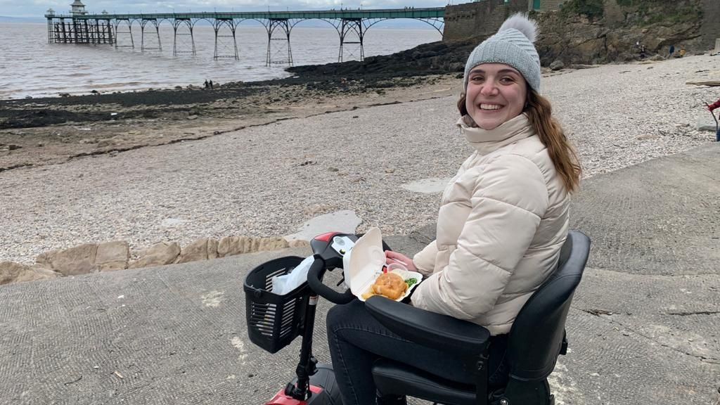 Chloe Boobier is sat in a wheelchair in front of the sea, with a box of what appears to be fish and chips on her lap. She is wearing a woolly hat and a white coat.