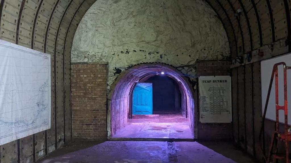 A large underground room with an arched ceiling which leads into a tunnel lit by a purple light.