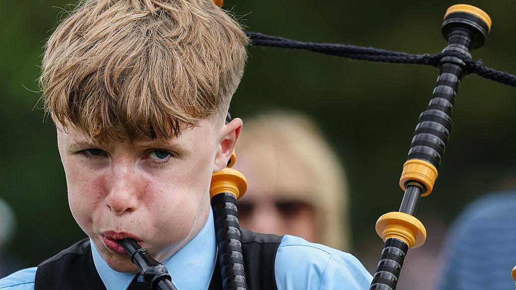 A close-up of a boy who is blowing his cheeks out as he plays on bagpipes. His eyes are half closed.