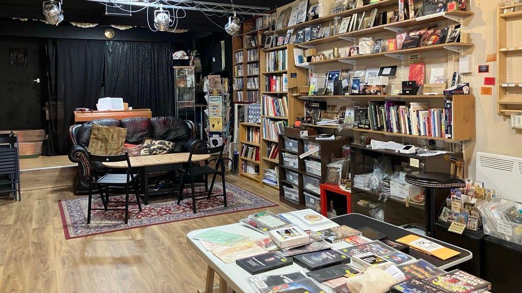 Interior of a cozy shop with wooden floors, shelves filled with books and mystical items, a table displaying cards and pamphlets, and a seating area with a sofa and chairs in front of a black curtain.