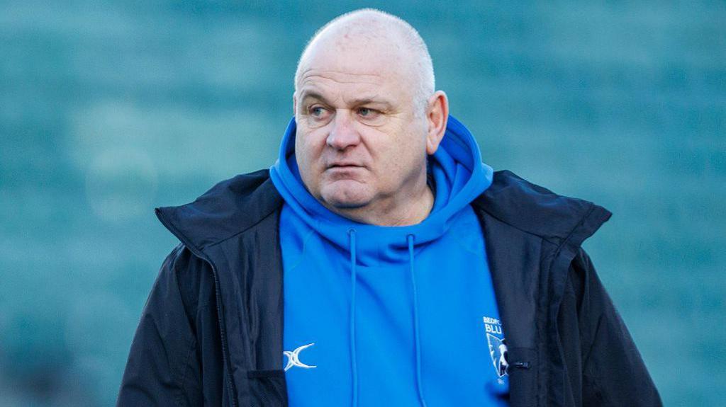 A serious looking Mike Rayer wearing a dark blue coat over a light blue Bedford Rugby Club hoodie