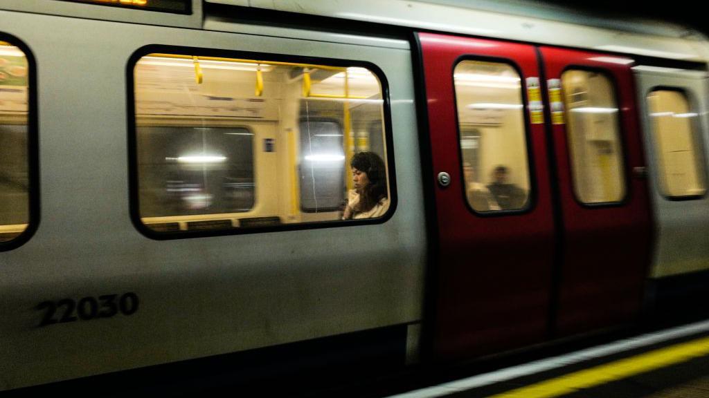 A London Tube train going through a station. The lights inside the train are yellow and a woman with dark hair is sitting alone next to the window. Two other passengers can be seen further down the carriage but they are blurred with the speed of the train.