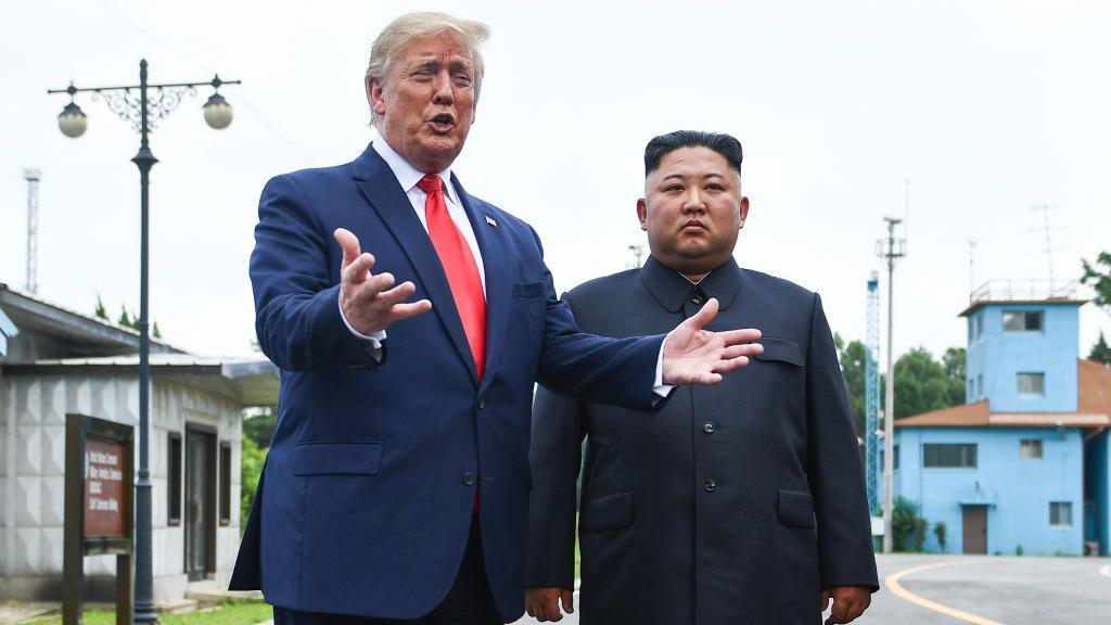 Standing on a road in the Joint Security Area, Trump gestures with his hands as he speaks, wearing a navy suit and red tie. Beside him stands Kim Jong Un, wearing a black button-up suit, looking solemn with his hands by his side.