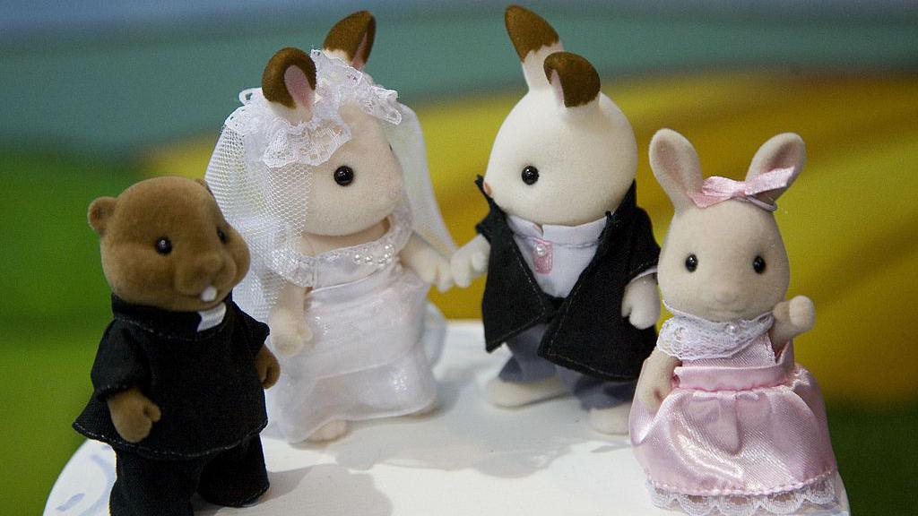 Four Sylvanian Families dolls are stood on what appears to be a wedding cake. There is a beaver-type toy dressed as a priest on the far left, second from left is a rabbit wearing a wedding dress, on her left is another rabbit dressed as a groom, then on the furthest right there's a smaller rabbit dressed in what appears to be a bridesmaids dress. It's pink, and she's wearing a matching pink ribbon on her head.