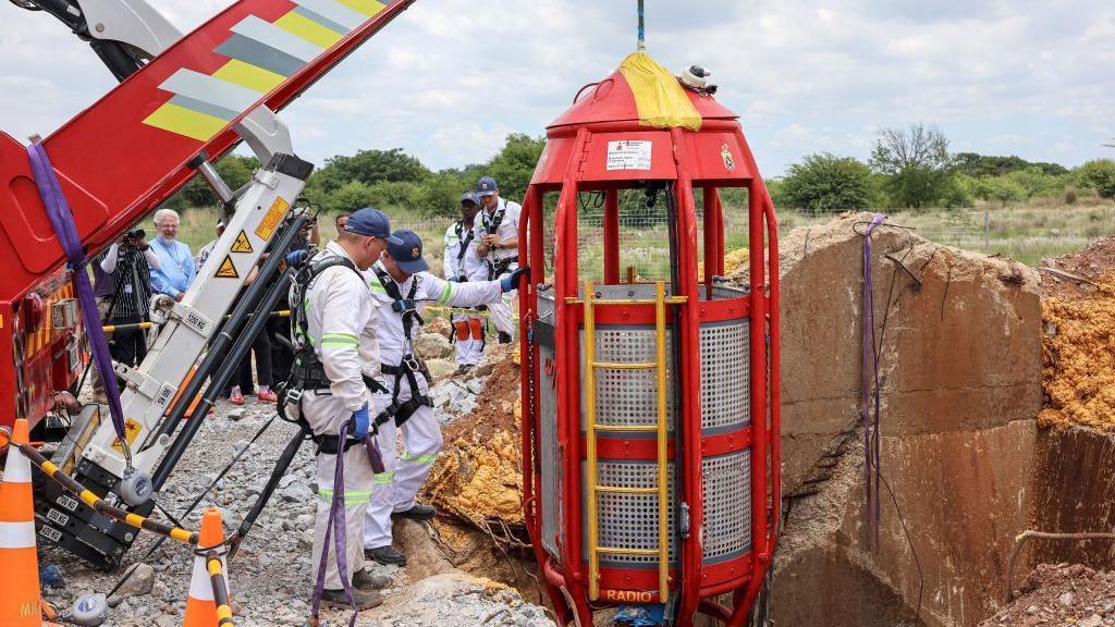 Two men dressed in white boiler suits stand next to a red metal cage attached to a hoist as they get it ready to be lowered down into a mine shaft.