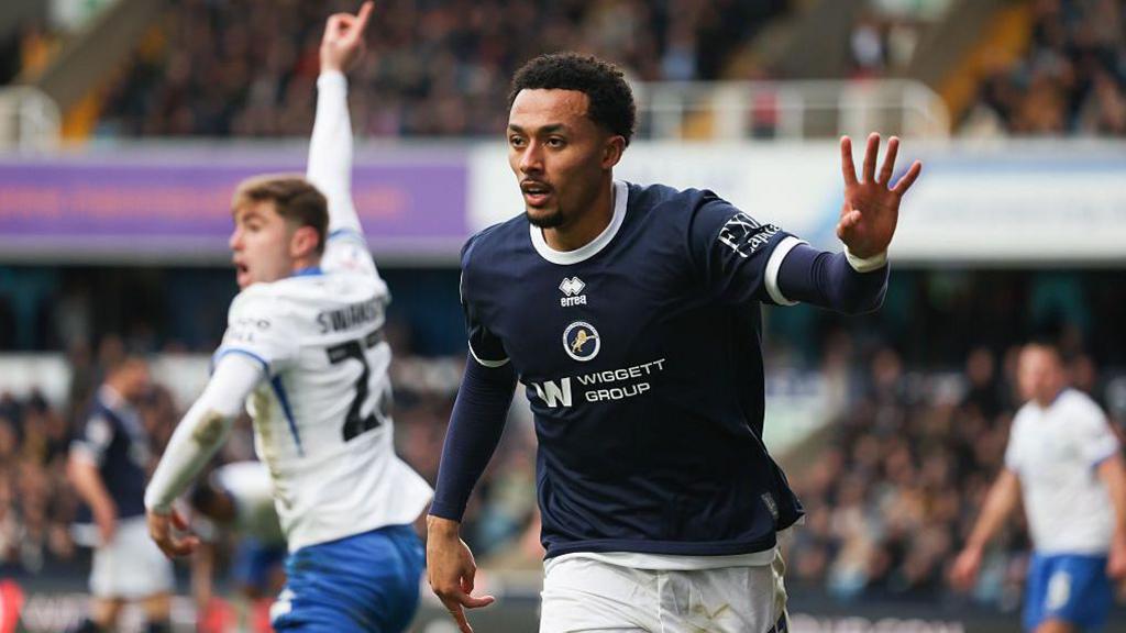 Millwall's Femi Azeez, wearing a dark blue shirt and white shorts, runs away with his left arm half raised after scoring a goal