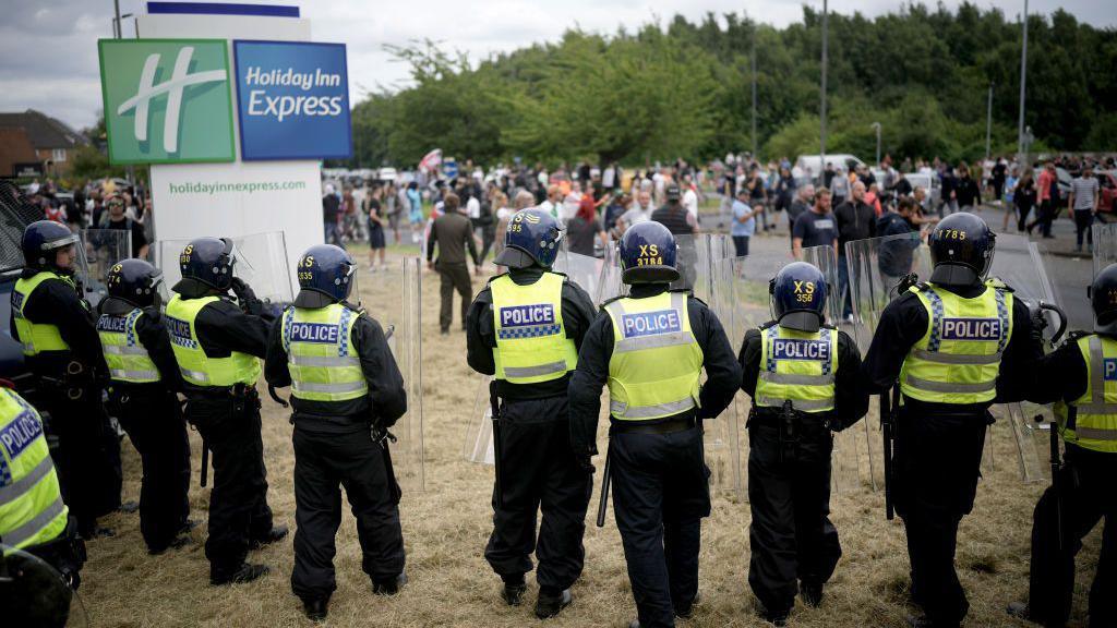 A line of police officers wearing yellow vests, some of them are holding riot shields and batons. There is a Holiday Inn Express sign in the background. There are a large number of people standing on the street behind the police officers.