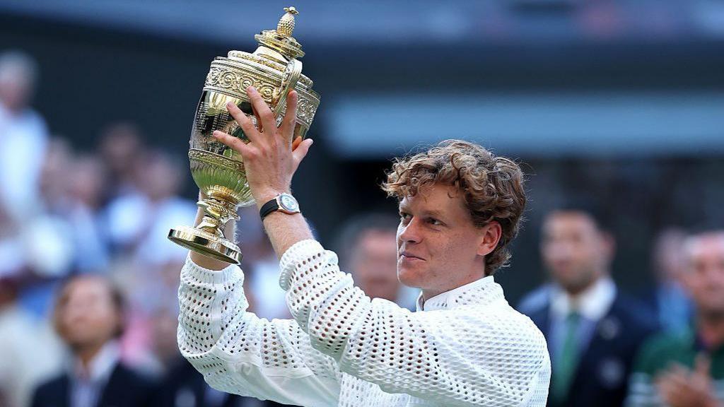 Jannik Sinner poses for a photo with The Gentlemen's Singles Trophy. He has curly light brown hair and is wearing a white jacket and a watch with a gold face and black strap. He is smiling and holding the gold trophy up above his head.