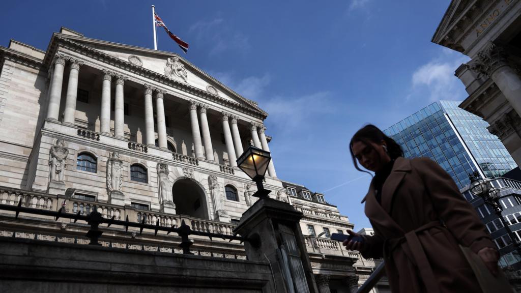 The Bank of England building with a blue sky behind it, in the foreground a woman stands on her phone