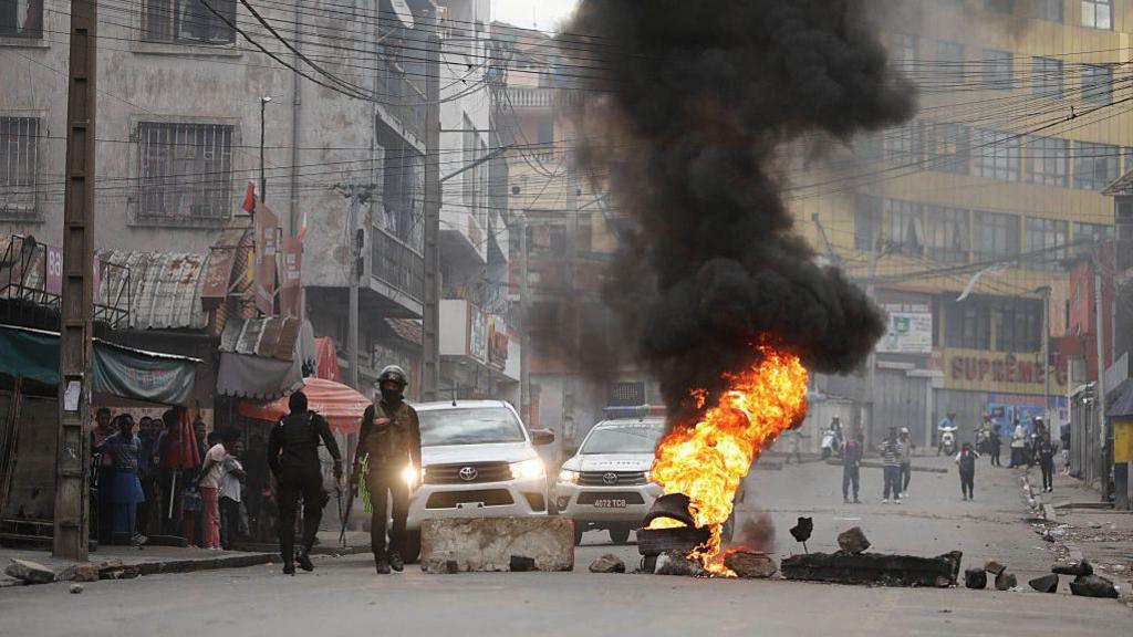 A wide shot of a main road with a burning barricade made up of tires and large concrete blocks. Security officials and their vehicles can be seen next to the barricade.
