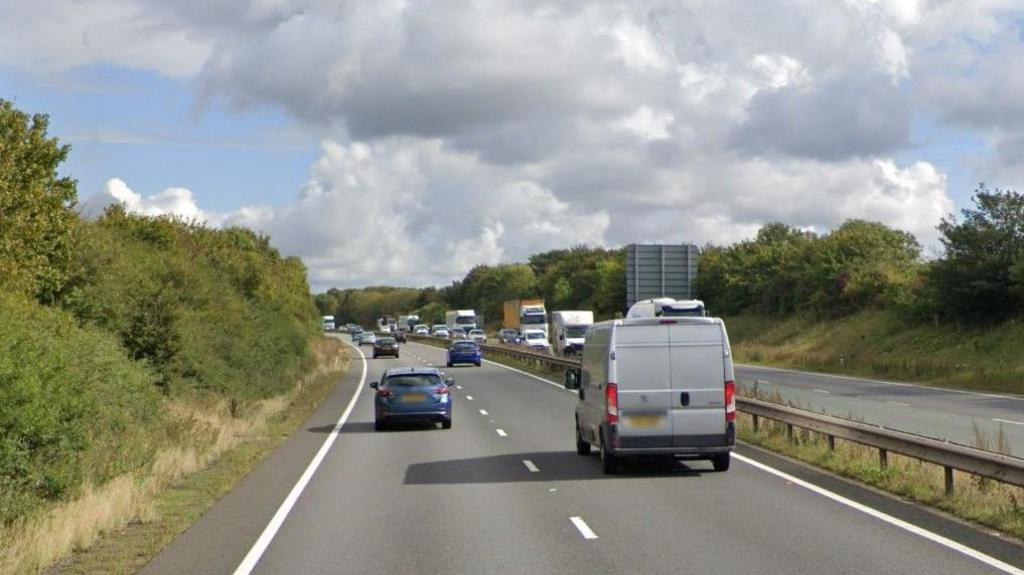 The A14, showing two dual carriageways, with vehicles on the road. A central reservation, a traffic sign in the distance, bushes and grass either side, a large sky, with grey clouds in the sky.
