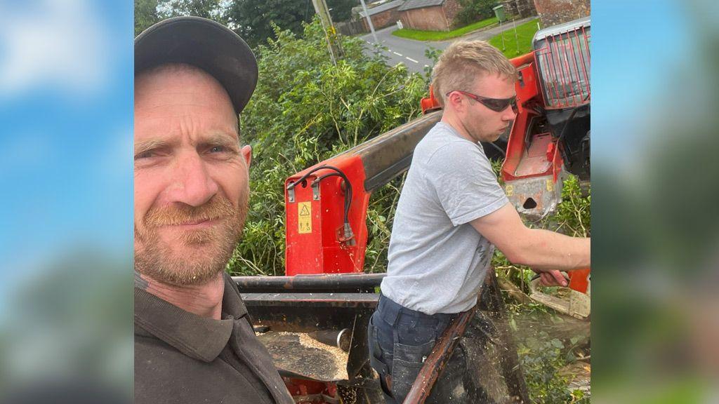 A selfie taken by Daniel Graham shows Daniel Graham in the foreground and Adam Carruthers working behind him. Daniel Graham is looking at the camera and wearing a black cap and black shirt. Adam Carruthers is side on and wearing sunglasses and a grey top
