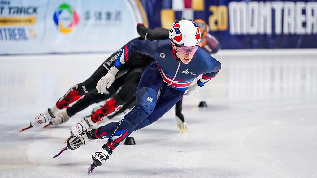 Niall Treacy competing in the men's 1,000m final. He is wearing a Great Britain skating uniform and wearing a white helment which also has red and blue markings.