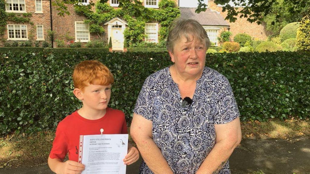 A woman stands beside a young boy, who holds up a petition form.