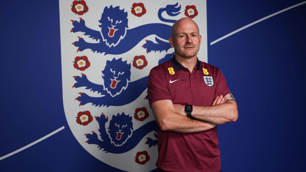 Lee Carsley standing in front of the three lions flag