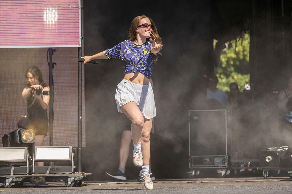 A woman in a tartan transmt football top dances on a stage weraing a short white skirt and sunglasses.
