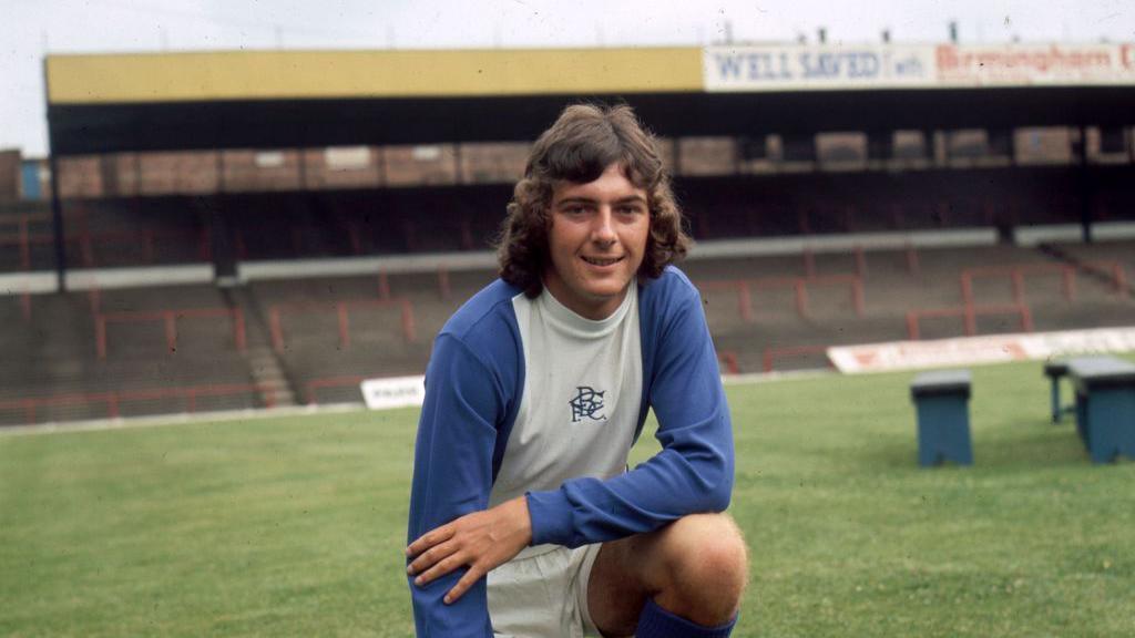Footballer Trevor Francis is photographed in an empty football ground in 1971. He is wearing blue and white football gear and is kneeling down on the grass.