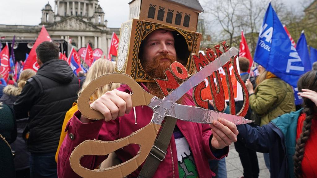 A man holds a cardboard placard that reads: NO MORE CUTS