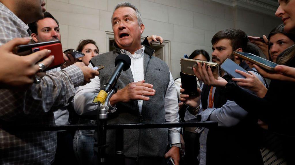 Don Bacon, wearing a grey sweater vest over a white dress shirt, gestures as he answers questions surrounded by a gaggle of Capitol Hill reporters aiming microphones and phones in his direction