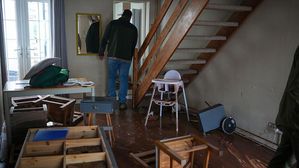 Tu mewn i gartref un o drigolion Trefynwy. Mae'r dodrefn wedi'u troi ar ben eu waered, a'r llawr yn wlyb wedi'i ddifetha. Mae dyn a'i gefn atom yn sefyll ger gorddrws y ty