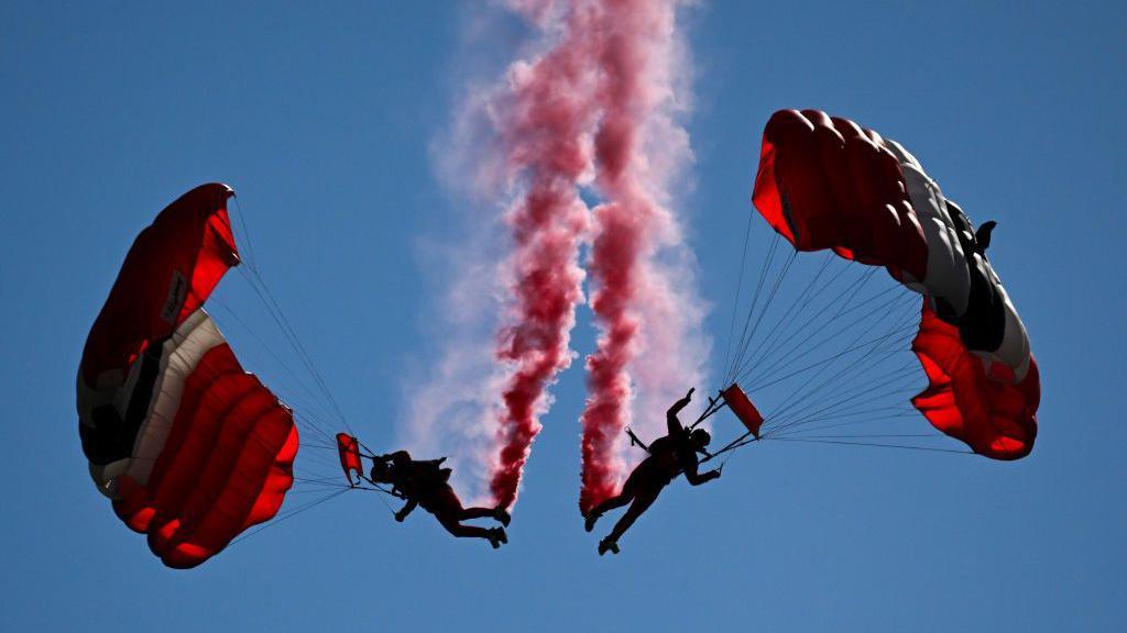 Two people in the sky with red parachutes. They are close to each other, with a red trail of smoke coming from their feet.