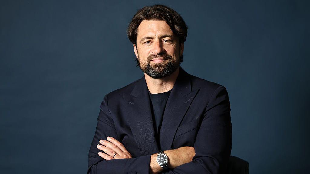 A smiling Russell Martin with his arms folded and wearing a dark blue suit jacket and dark blue t-shirt