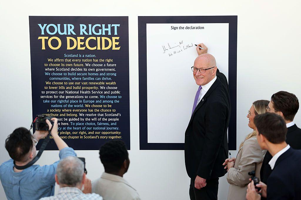 John Swinney - a bald man with glasses in a dark suit - smiles as he signs a declaration about "your right to decide" at a campaign event promoting Scottish independence