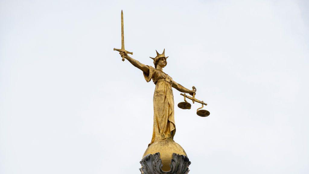 Gold Lady of Justice statue with a sword in one hand and scales in the other. She is wearing a crown.