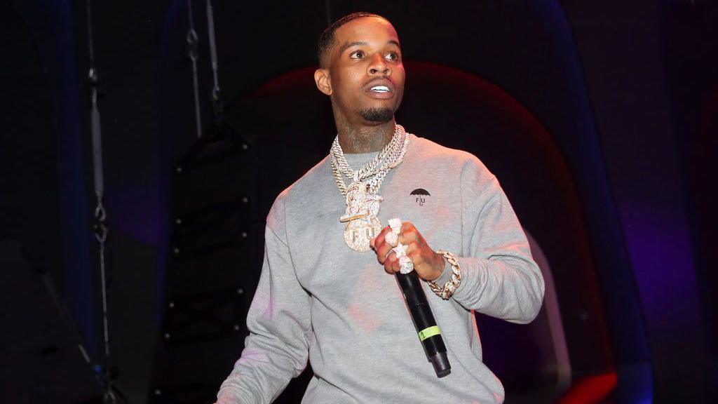 Rapper Tory Lanez is seen on stage holding a microphone. He is wearing a gray sweatshirt and multiple diamond necklaces and a big diamond ring on each finger.