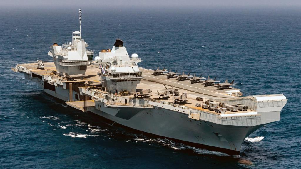 A large grey aircraft carrier in a still, blue sea with a light blue sky.