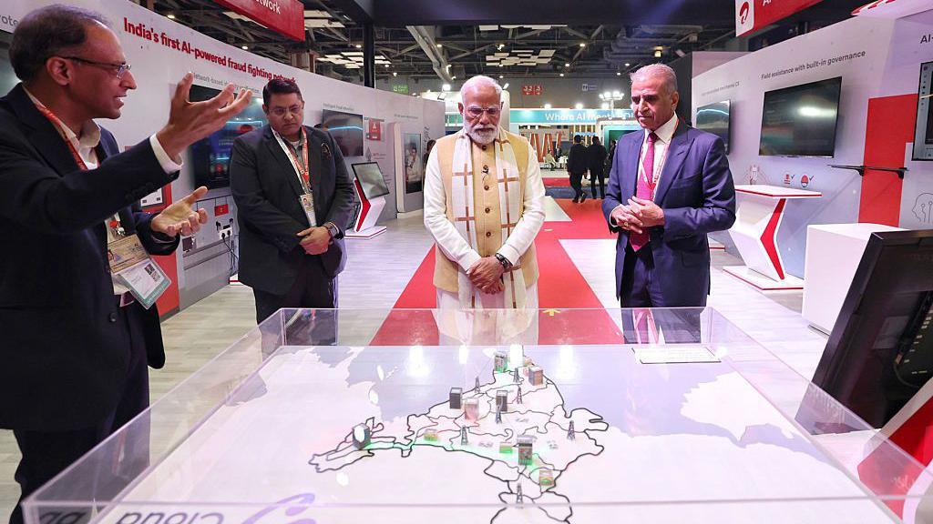 Prime Minister Narendra Modi seen wearing a white kurta and beige vest flanked by three men in suits at the India AI Impact Summit 2026. He is standing before a map of India shown under a glass case.