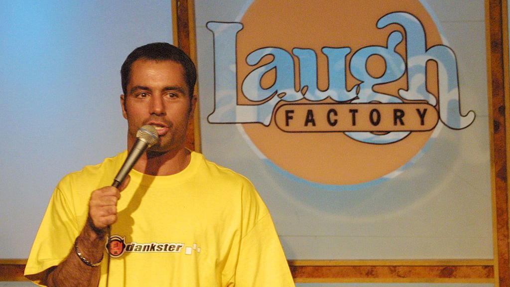 Joe Rogan, wearing a yellow shirt with a black logo that reads 'dankster', speaks holding a microphone during a stand-up set at the Laugh Factory in Hollywood in 2001