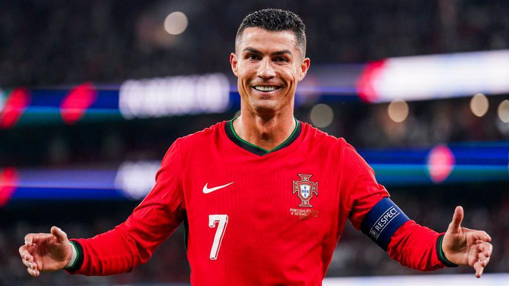 Cristiano Ronaldo, in a bright red Portugal kit and wearing the captain's armband, celebrates scoring with his arms wide and a big grin on his face.