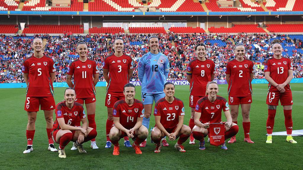 Llun o dîm merched Cymru mewn rhes ar y cae cyn chwarae