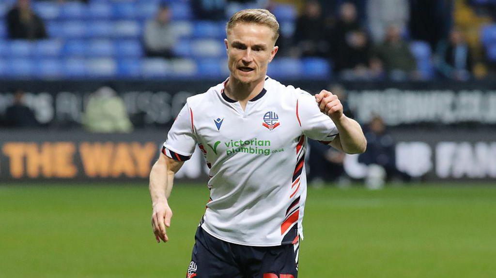 Kyle Dempsey in action for Bolton Wanderers. He is wearing a white home shirt and navy blue shorts. His left arm is raised as he prepares to kick the ball, which is out of view.