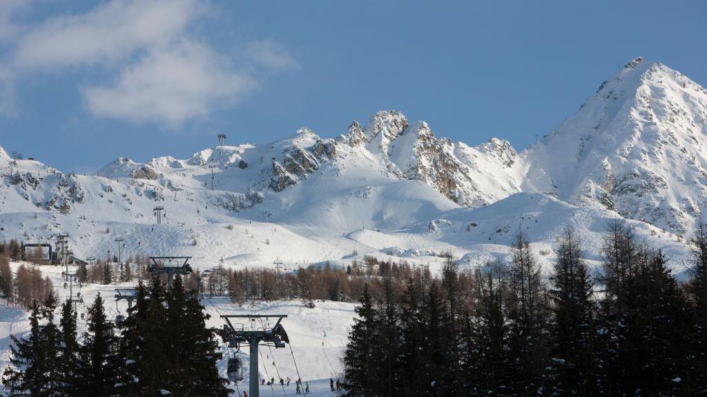 Snow-covered mountains under a light blue sky. Tall trees and a ski lift can be seen in the foreground.