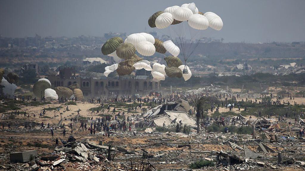 Several parachutes with aid packages attached fall onto a barren landscape littered with ruined buildings. Hundreds of people move towards the aid packages. 