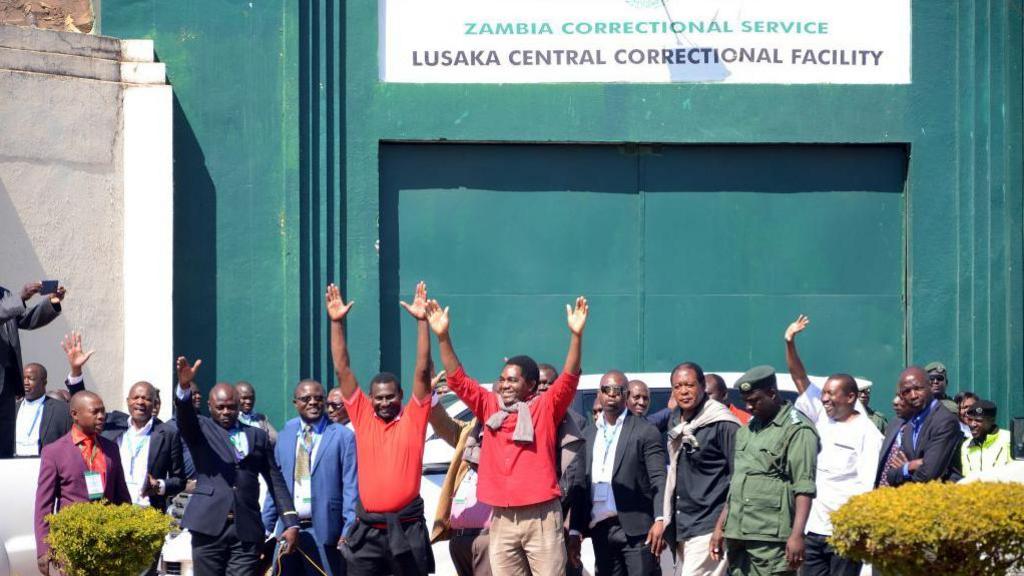 A group of people with their arms raised stand in front of a green gate with the sign Lusaka Central Correctional Facility above.