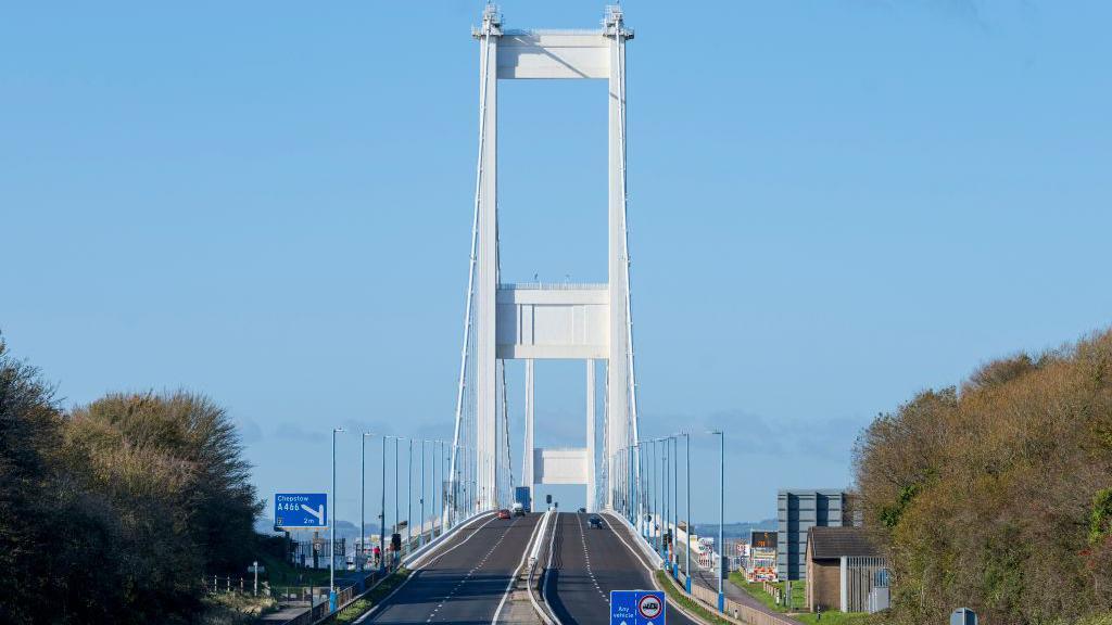 A shot of the Severn Bridge, looking from the English side with a sign showing a junction for Chepstow in view to the left. Trees flank the bridge at either side.