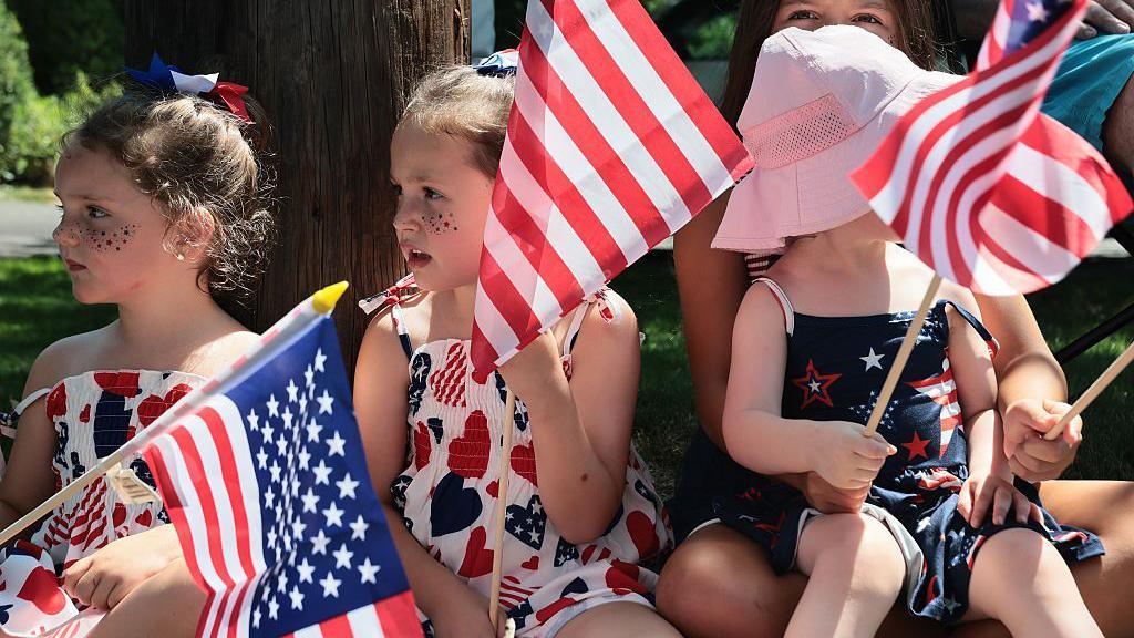 Three young girls sitting down and holding US flags, watching an event. They are all dressed in red, blue and white clothes.
