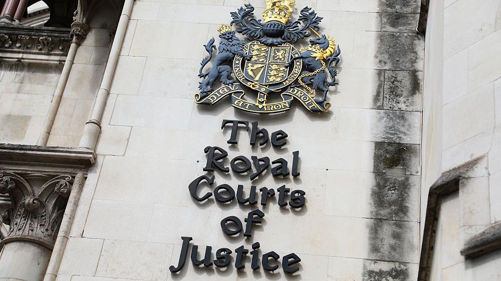 A view of the Royal Courts of Justice, showing a close -up of the name of the court beneath a heraldic shield on the outside of the building