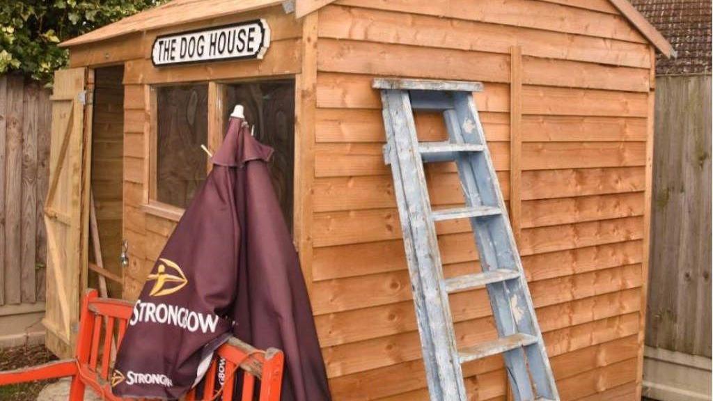 A small wooden garden shed with a sign saying The Dog House on it. There is a step ladder leaning against one side, with a wooden bench and garden umbrella at the front of the shed.