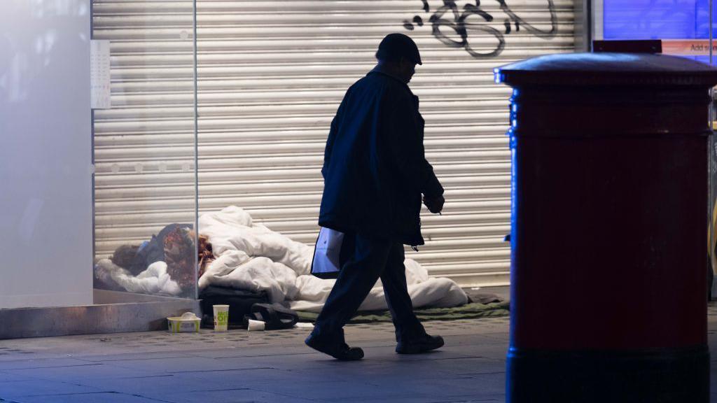 A man sleeps under a white duvet in front of a closed shop front. A man is walking past them and a post box can be seen in shadow to the right of them