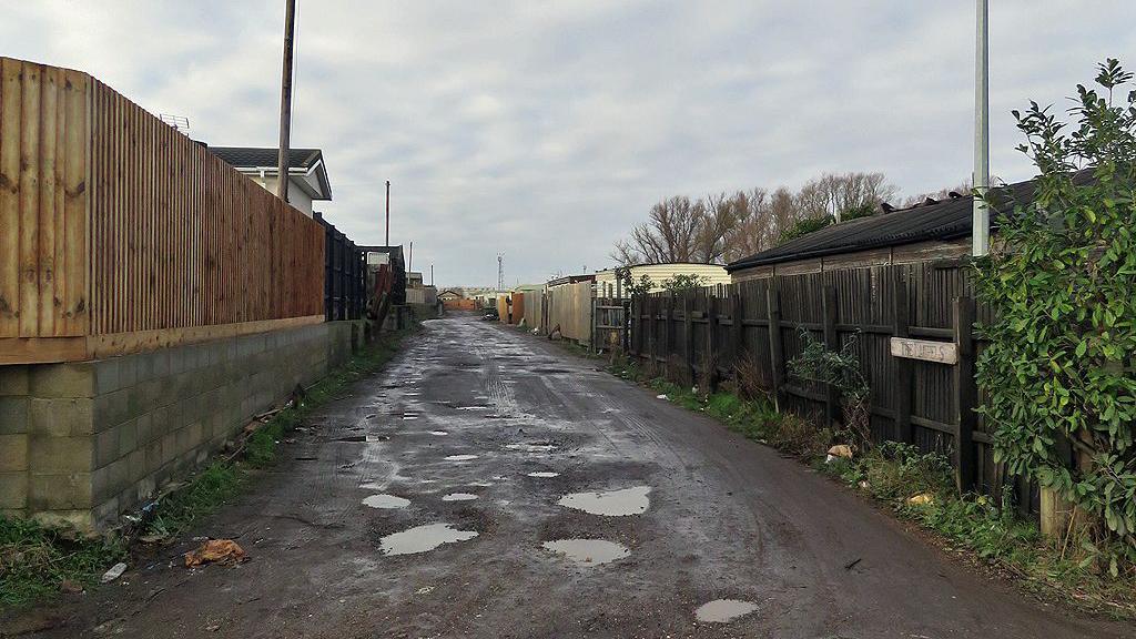 A muddy path with puddles has wooden fencing on either side as well as a white sign that says 'The Laurels'. The tops of mobile homes can be seen above the fences.