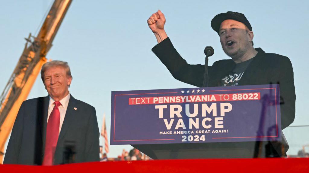 Donald Trump and Elon Musk on-stage at a Trump-Vance campaign event in 2024. Musk is speaking at a podium bearing the campaign logo, while Trump stands next to him, smiling.