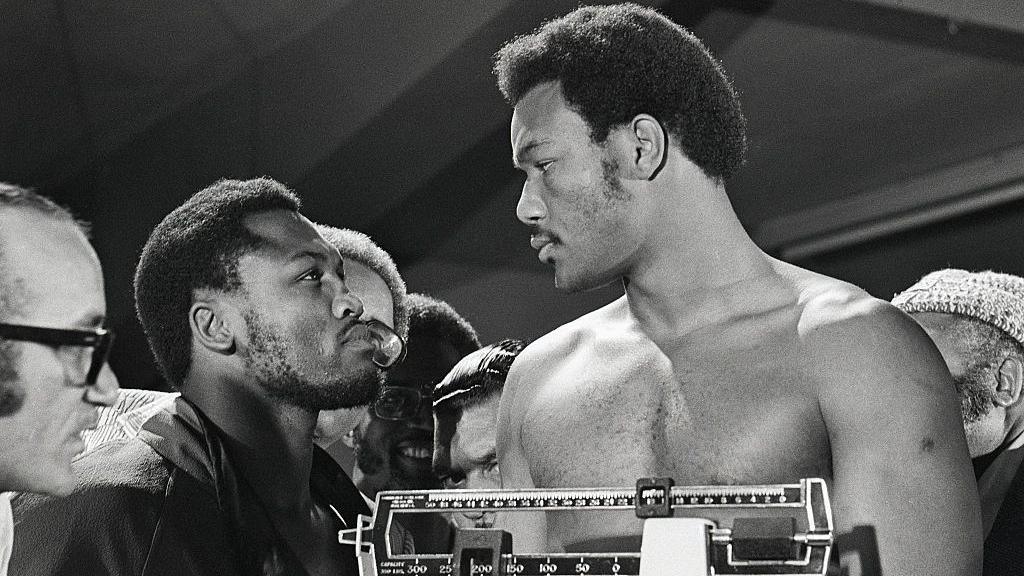 Joe Frazier stares at George Foreman, who is standing on the scales